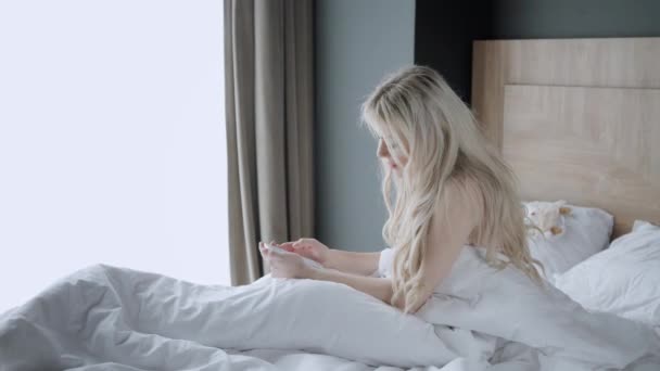 Usa o telefone. Verifica e-mail e mensagens de mídia social. A jovem acorda, senta-se numa cama confortável. Manhã no quarto de hotel . — Vídeo de Stock