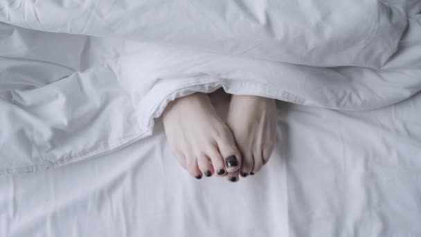 Jonge vrouwen voeten onder de witte deken. Concept voor gezonde zorg. Ochtend in hotelkamer. — Stockvideo