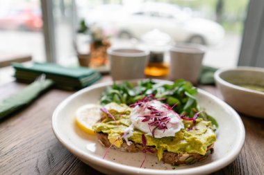 Kafede sabah, meşe masada. Avokado ile kepekli ekmek tost ile sağlıklı kahvaltı, yeşil salata ile haşlanmış yumurta. Arka Planda Yeşil çay.
