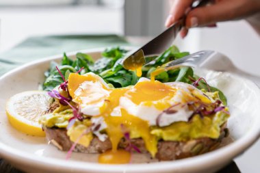 Genç bir kadın kahvaltısını çatal bıçakla yer, sabah kafede. Sağlıklı kahvaltı, tam ekmekli tost ve avokado, haşlanmış yumurta ve yeşil salata.