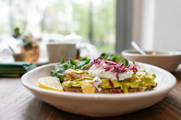 Morning in cafe, oak table. Healthy breakfast with wholemeal bread toast with avocado, poached egg with green salad. Green tea on the Background.