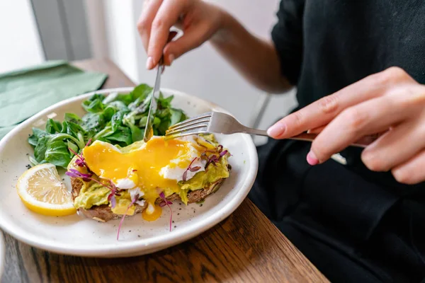 Young woman eat breakfast with knife and fork, Morning in cafe. Healthy breakfast with wholemeal bread toast with avocado, poached egg with green salad — 스톡 사진