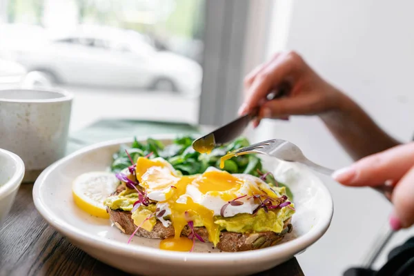 Young woman eat breakfast with knife and fork, Morning in cafe. Healthy breakfast with wholemeal bread toast with avocado, poached egg with green salad — 스톡 사진