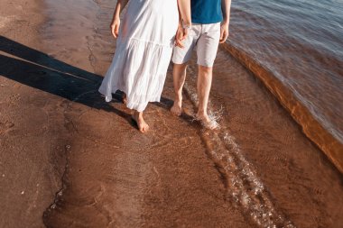 Ayaklar suda yaz. Genç yalınayak çift sahil boyunca yürüyor. Aşk hikayesi. Dalgalar ve kum. Dinlenme kavramı.