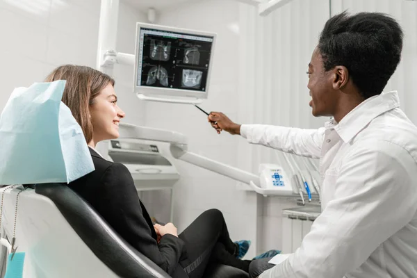 Un joven dentista africano muestra rayos X en el monitor. Mujer en la silla del dentista en la clínica dental. Medicina, salud, concepto estomatológico. dentista tratando a un paciente . — Foto de Stock