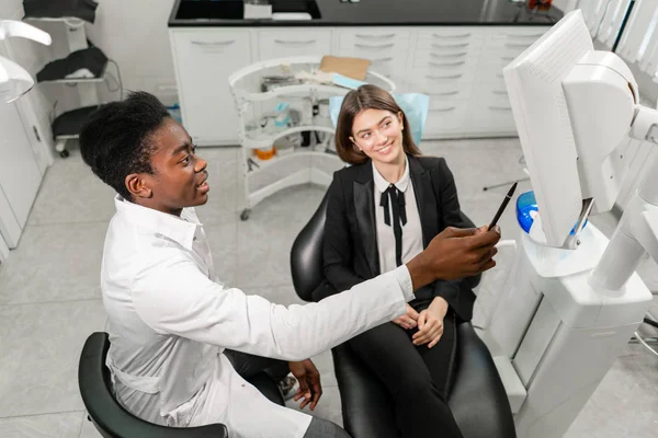 Um jovem dentista africano mostra raios-X no monitor. Mulher na cadeira de dentista na clínica dentária. Medicina, saúde, conceito de estomatologia. dentista tratando um paciente . — Fotografia de Stock