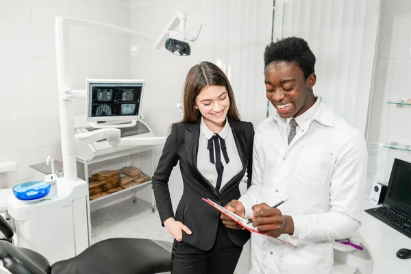 Ženská pacientka na zubní klinice. Mladý africký zubař dělá diagnózu a zaznamenává doporučení. Koncepce medicíny, zdraví, stomatologie. zubař ošetřující pacienta. — Stock fotografie