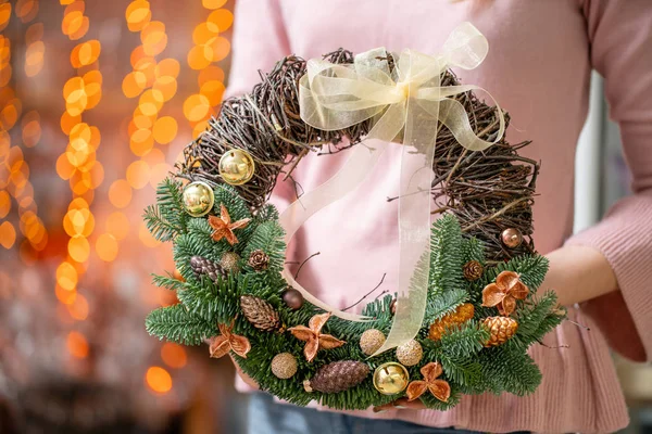 Bella corona festiva di abete rosso fresco con giocattoli a sfera nelle mani della donna. L'umore natalizio. Bokeh di ghirlanda luci su sfondo . — Foto Stock