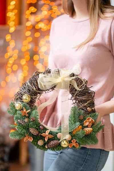 Όμορφο εορταστικό στεφάνι από φρέσκο έλατο με παιχνίδια μπάλα σε γυναικεία χέρια. Χριστουγεννιάτικη διάθεση. Τα φώτα του μπόκεη του Γκάρλαντ στο παρασκήνιο. — Φωτογραφία Αρχείου