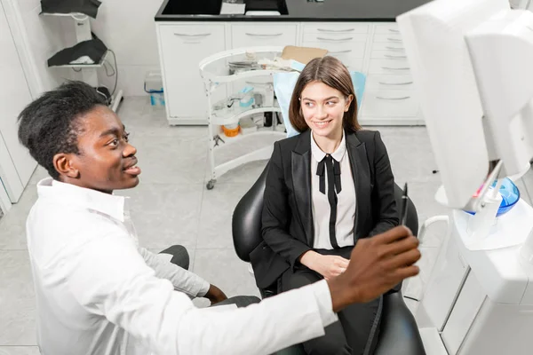 Un joven dentista africano muestra rayos X en el monitor. Mujer en la silla del dentista en la clínica dental. Medicina, salud, concepto estomatológico. dentista tratando a un paciente . —  Fotos de Stock