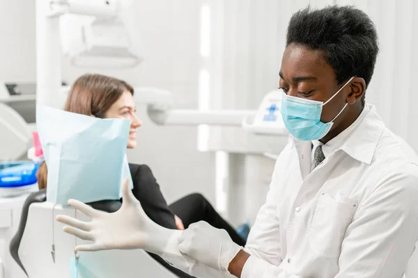 El doctor se pone guantes de goma. Joven dentista africano con un paciente. Mujer en la silla del dentista en la clínica dental. Medicina, salud, concepto estomatológico. dentista tratando a un paciente . — Foto de Stock