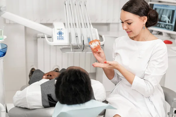 Mujer joven dentista que trata los conductos radiculares en la clínica dental. Joven afroamericano macho con mal diente acostado en silla de dentista con boca abierta . — Foto de Stock