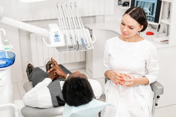 Mladá žena zubař ošetřující hlavní kanály na zubní klinice. Mladý Afričan americký muž se špatnými zuby ležet na židli zubaře s otevřenou pusou. — Stock fotografie