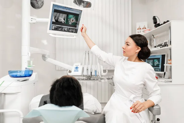 Mujer joven dentista que trata los conductos radiculares en la clínica dental. Joven afroamericano macho con mal diente acostado en silla de dentista con boca abierta . — Foto de Stock