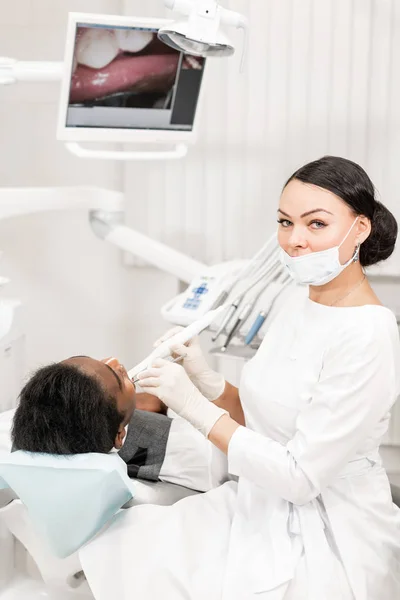 Inspección con una macro cámara dental y muestra el resultado en el monitor. Mujer joven dentista que trata los conductos radiculares en la clínica dental. Hombre paciente acostado en silla de dentista con la boca abierta . — Foto de Stock