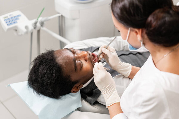 Молодая женщина-дантист лечит корневые каналы в стоматологической клинике. Молодой афроамериканец с плохими зубами лежит на стуле стоматолога с открытым ртом
.