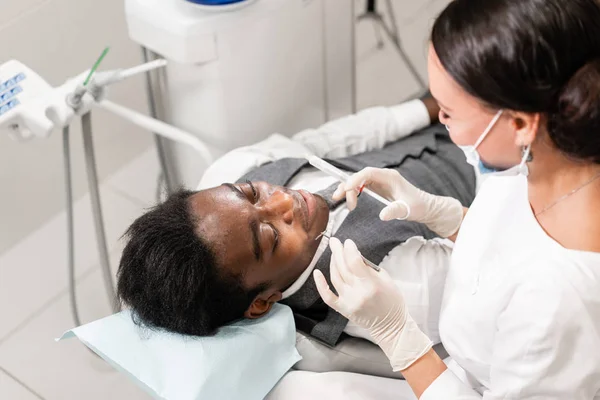 Mladá žena zubař ošetřující hlavní kanály na zubní klinice. Mladý Afričan americký muž se špatnými zuby ležet na židli zubaře s otevřenou pusou. — Stock fotografie