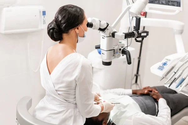 Mladá žena zubař léčení kořenových kanálků pomocí mikroskopu v zubní klinice. Muž pacient ležící na zubní křeslo s otevřenými ústy. Lékařství, zubní lékařství a zdravotnictví koncepce. Stomatologické zařízení — Stock fotografie