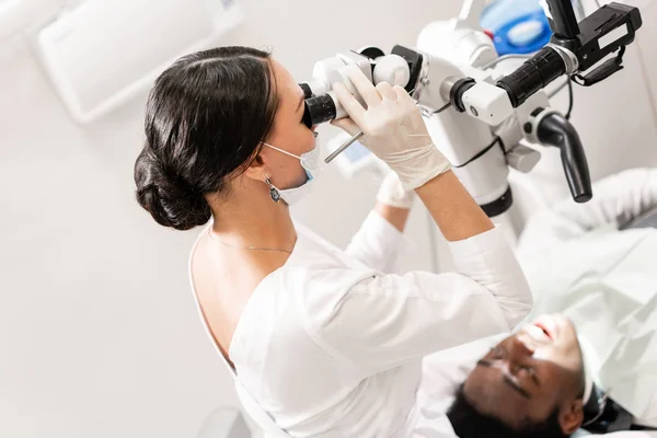 Jonge vrouw tandarts behandeling van root grachten met behulp van de Microscoop in de tandheelkundige kliniek. Man patiënt liggend op een tandarts stoel met open mond. Geneeskunde, tandheelkunde en gezondheidszorg concept. Tandheelkundige apparatuur — Stockfoto