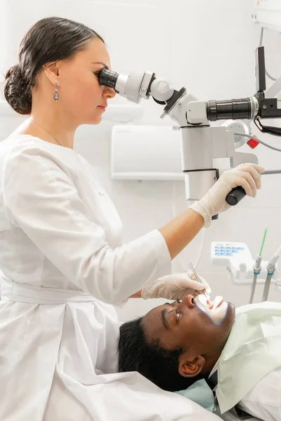 Mikroskop moderního vybavení v zubní ordinální kanceláři. Mladá žena zubař ošetřující kořenové kanály. Pacientka leží na židli zubaře s otevřenou pusou. Koncepce medicíny, stomatologie a zdravotní péče. — Stock fotografie