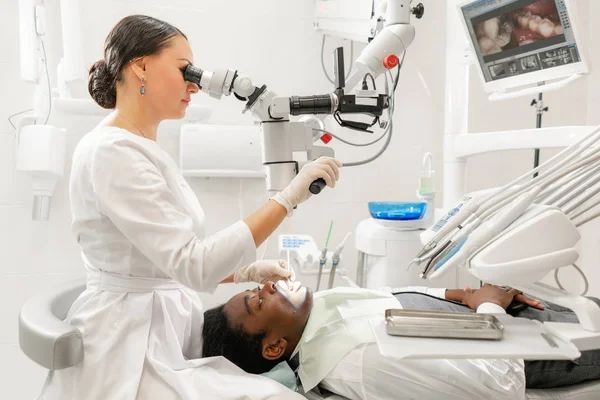 Mujer joven dentista que trata los conductos radiculares con microscopio en la clínica dental. Paciente hombre acostado en silla de dentista con la boca abierta. Concepto de medicina, odontología y salud. Equipos dentales —  Fotos de Stock