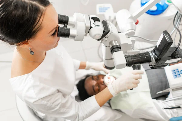 Microscopio de equipo moderno en consultorio dental. Mujer joven dentista tratando conductos radiculares. Paciente hombre acostado en silla de dentista con la boca abierta. Concepto de medicina, odontología y salud . —  Fotos de Stock