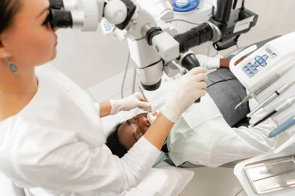 Microscopio de equipo moderno en consultorio dental. Mujer joven dentista tratando conductos radiculares. Paciente hombre acostado en silla de dentista con la boca abierta. Concepto de medicina, odontología y salud . — Foto de Stock