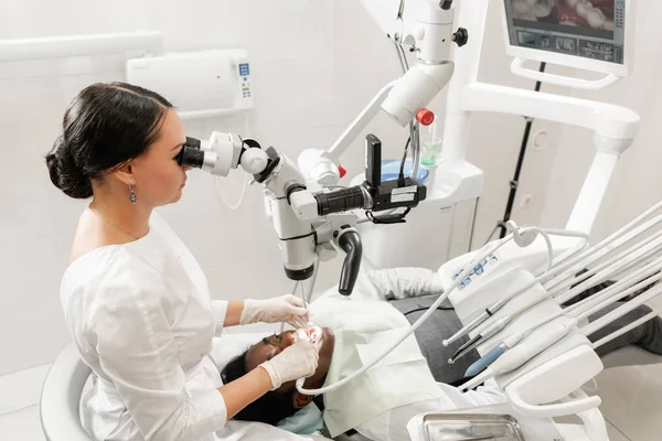 Microscopio de equipo moderno en consultorio dental. Mujer joven dentista tratando conductos radiculares. Paciente hombre acostado en silla de dentista con la boca abierta. Concepto de medicina, odontología y salud . —  Fotos de Stock