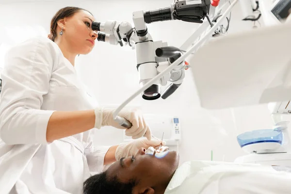 Microscopio de equipo moderno en consultorio dental. Mujer joven dentista tratando conductos radiculares. Paciente hombre acostado en silla de dentista con la boca abierta. Concepto de medicina, odontología y salud . — Foto de Stock