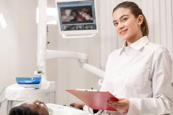 Encantadora doctora dentista hermosa. Joven paciente afroamericano en silla en la clínica dental. Medicina, salud, concepto estomatológico. dentista lleva a cabo la inspección y concluye. Mujer sonriendo — Foto de Stock