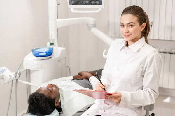 Encantadora doctora dentista hermosa. Joven paciente afroamericano en silla en la clínica dental. Medicina, salud, concepto estomatológico. dentista lleva a cabo la inspección y concluye. Mujer sonriendo — Foto de Stock