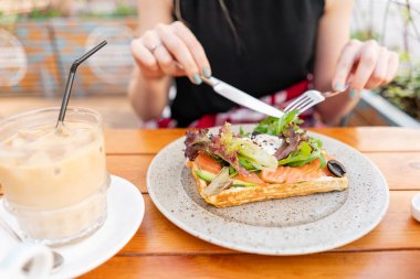 Somon, marul yaprakları ve haşlanmış yumurta ile geleneksel Belçikalı Waffle. Genç bir kadın bir yaz kafede kahvaltı yapıyor, bir çatal ve bir bıçak yakın çekim ile eller. Buz küpleri ile soğuk kahve