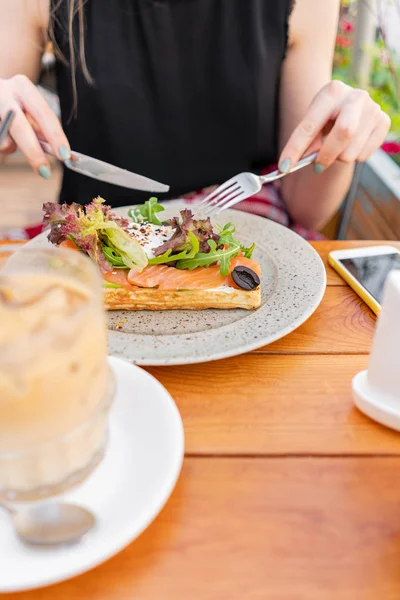 Tradiční belgická vafle s lososem, hlávkovým salátem a vajíčkem. Mladá žena snímá v letní kavárně, ruce s vidličkou a s nožem zblízka. Studená káva s ledem — Stock fotografie