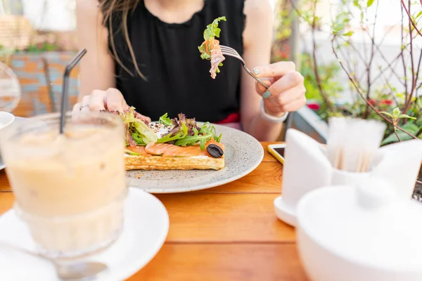 Traditional Belgian waffle with salmon, lettuce leaves and poached egg. A young woman is having Breakfast in a summer cafe, hands with a fork and a knife close-up. Cold coffee with ice cubes — Stock Photo, Image