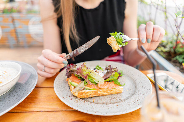 Traditional Belgian waffle with salmon, lettuce leaves and poached egg. A young woman is having Breakfast in a summer cafe, hands with a fork and a knife close-up. Cold coffee with ice cubes