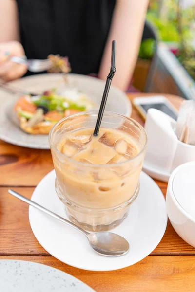 Focus in foreground on Cold coffee with ice cubes. A young woman is having Breakfast in a summer cafe. — Stock Photo, Image