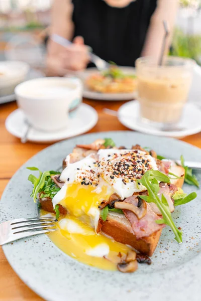 Breakfast and coffee in the summer cafe. Sandwich on a bun brioche with bacon, mushrooms, arugula and poached egg — Stock Photo, Image