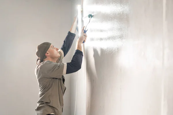 Young repairman smear on wall glue with a roller. . Worker glueing wallpapers on concrete wall. Repair the apartment. Home renovation concept. White Wallpaper for paint — 스톡 사진