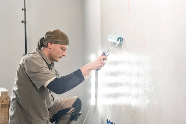 Young repairman smear on wall glue with a roller. . Worker glueing wallpapers on concrete wall. Repair the apartment. Home renovation concept. White Wallpaper for paint — 스톡 사진