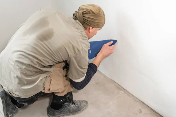 Молодий ремонтант з'єднується з пластиковим шпателем. Працівник приклеює шпалери на бетонну стіну. Ремонт квартири. Концепція ремонту будинку. Білі шпалери для фарби — стокове фото