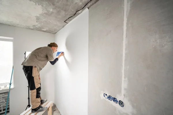 Fiatal műszerember igazítja a műanyag spatula. Munkás ragasztás tapéta-ra beton fal. Az apartman megjavítása. Lakásfelújítás koncepció. Fehér tapéta festéshez — Stock Fotó