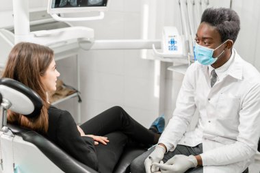 Genç Afrikalı erkek dişçinin hastası var. Diş kliniğinde dişçi koltuğunda oturan kadın. Tıp, sağlık, stomatoloji konsepti. bir hastayı tedavi eden dişçi.