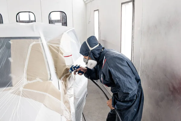 Car service station. Worker painting a white car in special garage, wearing costume and protective gear