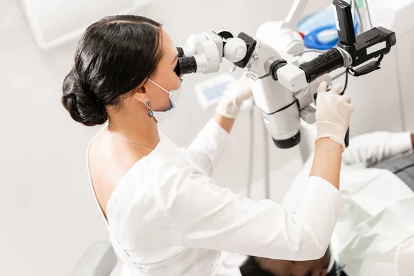 Молодая женщина-дантист лечит корневые каналы с помощью микроскопа в стоматологической клинике. Пациент лежит на стоматологическом стуле с открытым ртом. Медицина, стоматология и здравоохранение концепция. Стоматологическое оборудование — стоковое фото