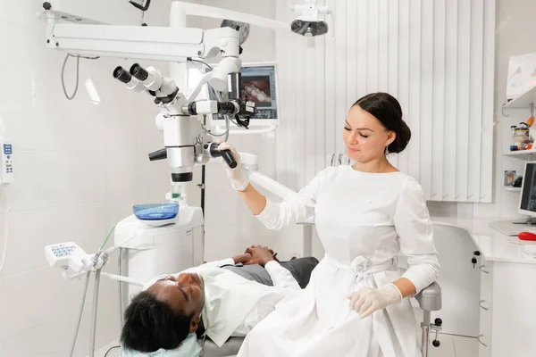 Portriat Mujer joven dentista con microscopio. Paciente hombre acostado en silla de dentista con la boca abierta. Concepto de medicina, odontología y salud . —  Fotos de Stock