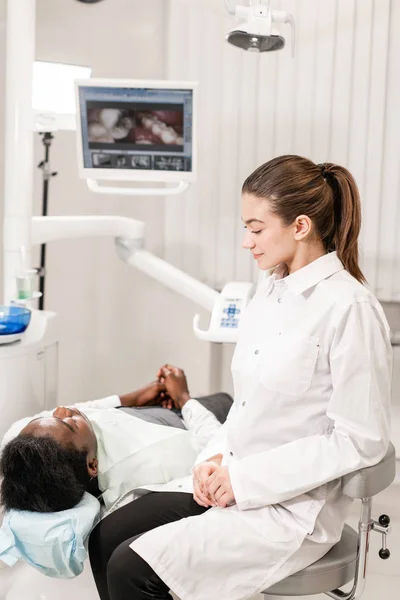 Encantadora doctora dentista hermosa. Joven paciente afroamericano en silla en la clínica dental. Medicina, salud, concepto estomatológico. dentista lleva a cabo la inspección y concluye. Mujer sonriendo — Foto de Stock