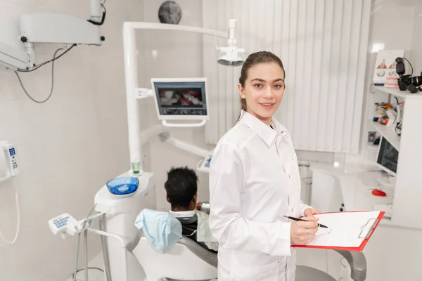 Encantadora linda menina dentista olha para a câmera. Jovem afro-americano na cadeira da clínica dentária. Medicina, saúde, conceito de estomatologia. dentista realiza inspeção e conclui . — Fotografia de Stock