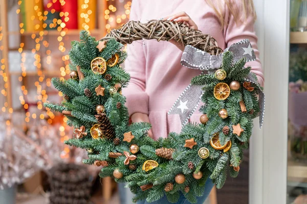Hermosa corona festiva de abeto fresco con juguetes de pelota en manos de mujer. Humor de Navidad. Bokeh de luces de guirnalda en el fondo . — Foto de Stock