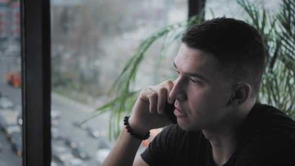 El joven habla por teléfono. Freelancer trabaja en netbook en coworking moderno. Programador en el trabajo remoto. Gente exitosa, hombre de negocios en una cafetería cómoda. Ventanas grandes altas — Vídeos de Stock