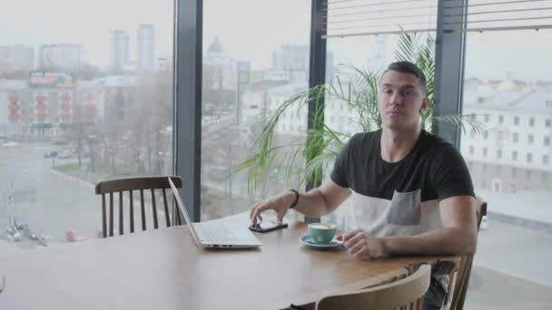 El joven habla por teléfono. Freelancer trabaja en netbook en coworking moderno. Programador en el trabajo remoto. Gente exitosa, hombre de negocios en una cafetería cómoda. Ventanas grandes altas — Vídeo de stock